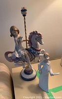 Two porcelain figurines displayed on table: Lladro carousel horse with seated girl and separate white stylized girl figurine holding tray