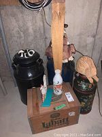 View of two large black metal milk cans, a wooden 'John Labatt Classic' crate, ceramic rooster on green base, white bottle with bull design, and floral vase with broom attached