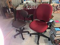 Photo of both office chairs, one red with armrests and one gray with no armrests, on concrete floor near books and other household items