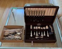 Overview of wooden casket of flatware and adjacent box of loose pieces on glass table