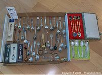 Overview of multiple silverplate souvenir spoons displayed on cardboard with some boxed demitasse spoon sets and a small group of cloisonné spoons on the side.