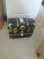Closed black retro trunk style chest with curved top, brown leather strap accents, and gold-tone metal hardware standing on a carpeted floor against a white door.