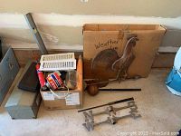 Overview of weather vane components, boxes with tools and hardware, the metal rooster piece, directional letters, and metal rods and spherical finial on the floor.