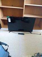 Front view of Samsung 31 inch flat screen TV resting on carpeted floor with wooden shelves in background. Power cord visible extending from the TV base.