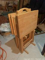 Side view of four light-colored wooden TV trays stacked on a wooden stand, showing the handle cutouts and tray surfaces.