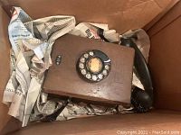 Top-down view of wooden rotary phone in a box cushioned with newspaper showing rotary dial and the phone resting horizontally.