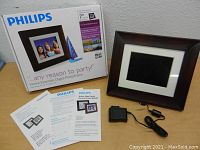 Philips digital photo frame displayed with box, power adapter, and manuals on a table against a white wall.
