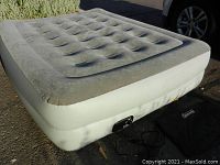 View of the inflated air mattress showing the gray top with air pockets and white sides, with the built-in pump unit visible along the bottom side and the carry bag beside it on the ground.