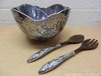 Front view of the large Lenox metal bowl with embossed dragonfly and floral designs, accompanied by two wood-handled utensils with metal floral pattern handles.