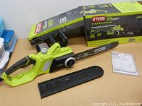 Ryobi electric chainsaw laid on a table with original box behind, blade cover beside chainsaw, and instruction manual visible.