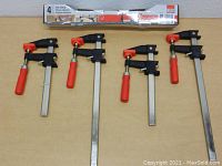 Four Bessey bar clamps laid horizontally on a wooden surface with box behind, displaying red handles and metal bars in different sizes.