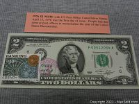 Photo showing front of the 1976 $2 bill with US Post Office cancellation stamp and 10-cent postage stamp, note placed on dark fabric background, including informational card about issue date.