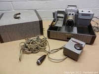 Vintage Bell & Howell TDC Selectron Headliner 303 slide projector placed inside an open carrying case with wired remote control and power cable arranged beside it.