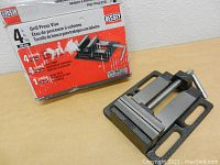 Photo showing the Bessey drill press vise with original red and white box in the background. Vise has black metal body, silver screw with handle, and mounting slots.