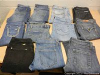 11 pairs of women's/girls jeans folded and arranged on a table in two rows, showing back pockets and colors including light blue, medium blue, dark blue and black.