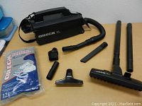 Complete Oreck XL vacuum cleaner with handle and shoulder strap. Accessories arranged on table including flexible hose, extension wands, various attachments, and vacuum bags.