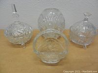 Clear crystal three-footed dishes with lids, round vase, and crystal basket vase arranged together on surface.