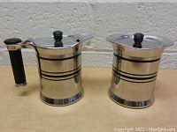 Front view of the two-piece vintage stainless steel cream and sugar set showing the handles, lids and decorative black stripes.