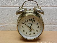 Frontal view of vintage brass Ingraham double bell alarm clock on peg legs showing white dial with black numerals and hands.