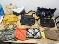 Top view of 10 various women's purses including metallic grey, mustard yellow, orange, black, floral embroidered, white perforated, beige, and patent black handbags and clutches.