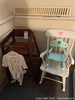Photo of three chairs: a brown wooden high chair, a large white kid-sized rocking chair with pastel accents, and a small blue doll rocking chair on the white chair seat. Also a Baby Dior baby outfit hanging nearby.