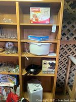 Wooden shelf holding various kitchen items including a white electric hot plate, beige slow cooker, white bread machine, black brownie maker pan and two cast iron pans. Boxed Pyrex measuring cups set also visible.