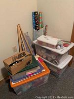 Stacked plastic bins contain assorted craft supplies including ribbons, gift wrap rolls, and gift bags near a wooden easel.