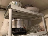 Photo showing a shelf with metal bakeware pans including a large bundt pan, along with plastic coffee carafe, pink plastic drinking glasses, and clear glass bowls.