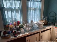 Wide shot of assortment of candles, glassware, ceramic serveware, and metal servers on counter under window with sheer curtains.