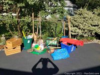 Wide view of garden tools, crates, bins, tarps, hose, and metal tins stacked on driveway with bushes in background