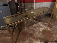 Full view of a glass and metal console table with tubular metal legs and clear rectangular glass top, placed on a concrete floor with some dirt and dust.