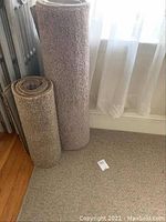 Rolled and laid flat tan tweed rugs near window and radiator, showing size and texture of rugs.