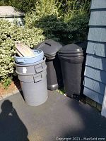 View of 5 trash cans of various sizes and colors: gray, blue, and black. Three black cans have lids and wheels. The gray can has a loose lid on top.