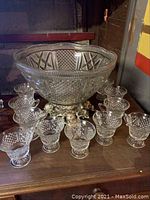 Full view of large glass punch bowl with metal footed base surrounded by 11 matching glass cups with footed bases