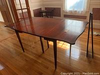 Vintage mahogany wood dining room table with four leaves partially extended, tapered legs, and drop side panels in a wood-floored dining room.