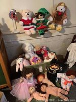 Wide view of lot with vintage dolls and assorted plush animals in a green bin and sitting nearby, including themed dressed bears and a bunny.