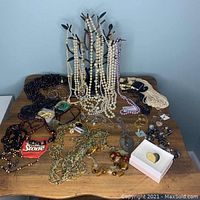 Photo showing multiple strands of faux pearl necklaces, beaded necklaces, pins, and rings on a wooden surface against a blue wall with black stand for hanging necklaces