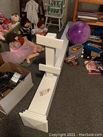 White wooden mailbox post lying on carpeted floor near dolls and boxes, showing overall structure including base and mounting arms.