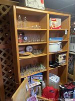 Front view showing two-column solid wood bookcase with multiple shelves filled with various household items for visual scale. The bookcase has a medium wood finish and solid construction.