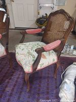 Front three-quarter view of one chair showing wooden frame, caned back, floral seat cushion and pink velvet arm pads