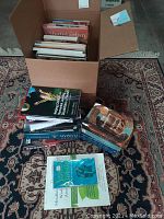 Box filled with multiple books of various sizes stacked inside a cardboard box and scattered on an Oriental style carpet. Visible titles include a vegetarian cookbook and assorted novels and educational books.