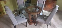 Photo showing the full dining set including the round glass top table with wooden carved base and four upholstered chairs around it.