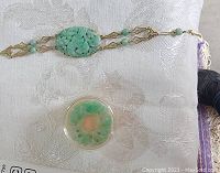 Photo of gold and jade bracelet with carved jade centerpiece and separate round jade container, displayed on white textured fabric.