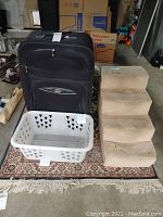 Photo showing white plastic laundry basket, beige carpeted pet stairs, and large black wheeled luggage placed on a carpet in a garage setting.
