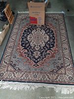 Full view of the rug showing the overall design, colors, and fringe, with a cardboard box partially on the rug.