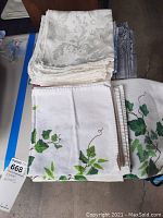 Image showing stacked folded white napkins with green ivy embroidery alongside cream damask fabric linens, placed on floor