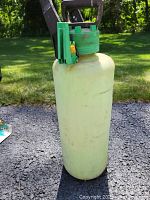 Side view of yellow 5 gallon sprayer tank with green pump handle, placed on gravel ground outside.