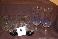 Set of six assorted drinking glasses on brown cloth background. Two beer mugs with handles, two tall pina colada glasses with blue design, two black bottom cocktail glasses.