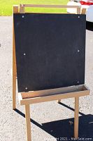 Wooden easel showing blackboard side with chalk tray attached along the bottom edge, viewed outdoors on pavement.