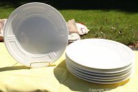 Six stacked white charger plates with Naxos pattern embossed design on rim, shown on yellow surface outdoors.